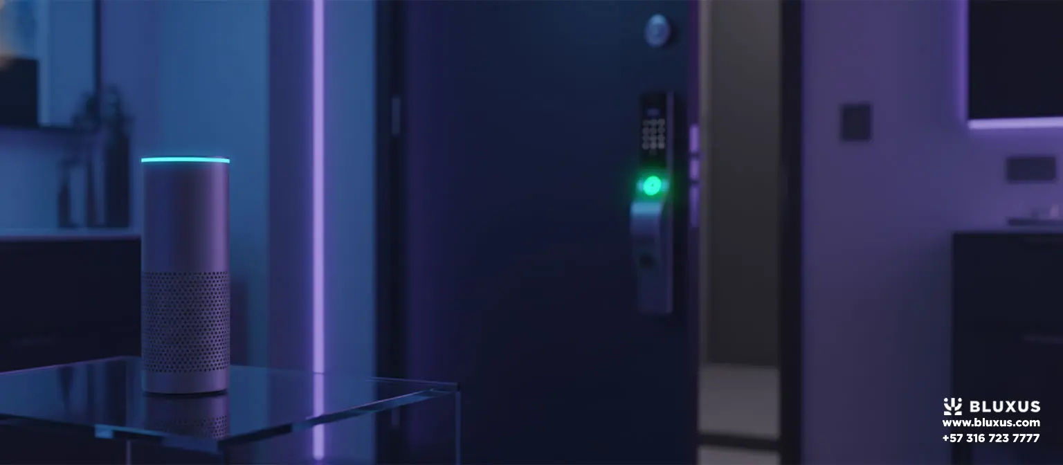 Altavoz cilíndrico iluminado con un anillo de luz cian sobre una mesa de cristal, con una puerta entreabierta al fondo que muestra el indicador verde de una cerradura electrónica en un ambiente nocturno.