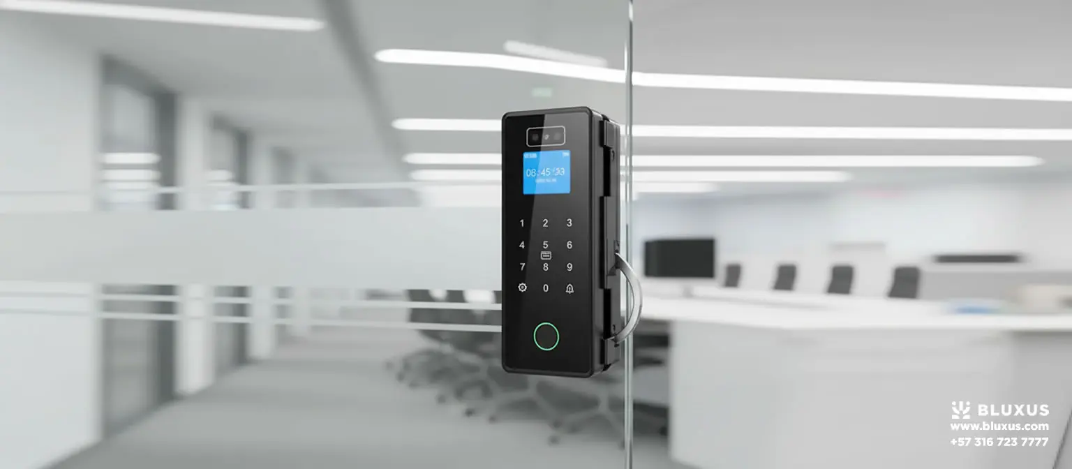 Mecanismo electrónico oscuro con pantalla azul iluminada, sujeto a presión sobre el panel de cristal transparente de una oficina moderna y brillante con escritorios al fondo.